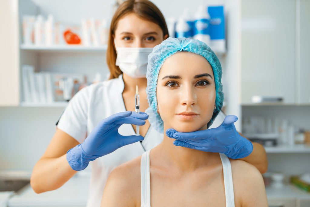 Cosmeticiană cu mască ține o seringă pregătită lângă fața unei paciente, procedură injectabilă