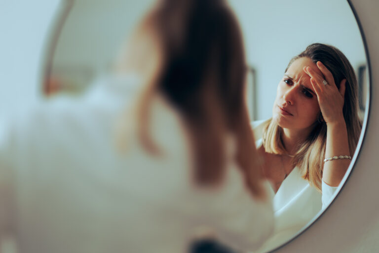 Femeie se privește îngrijorată în oglindă, examinându-și pielea frunții